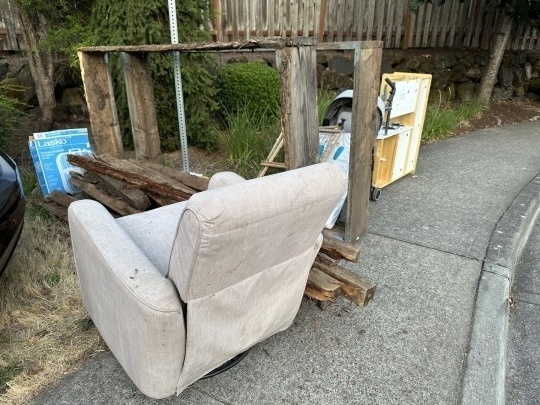 An old sofa, wooden beams, a dolly, and some other discarded items are placed on the side of a sidewalk near a wooden fence.