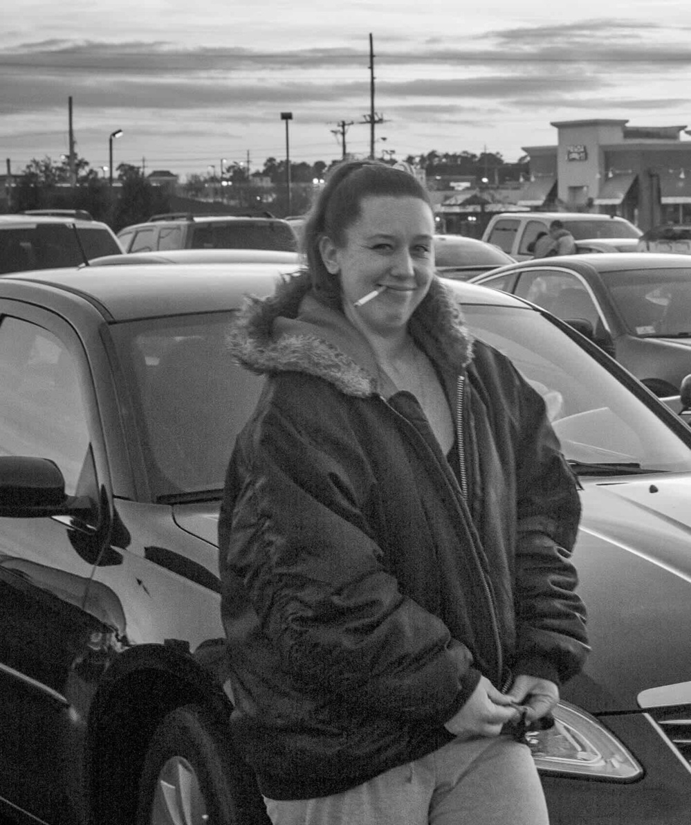 Person stands smiling with a cigarette in mouth wearing a large jacket in a busy parking lot near multiple parked cars under a cloudy sky with a shopping center in the background