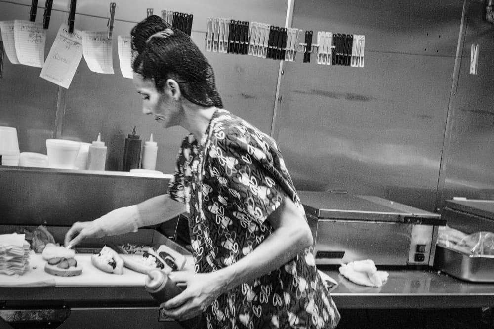 A person assembles food on a counter, surrounded by kitchen equipment and utensils, with order tickets clipped above and condiment bottles nearby.