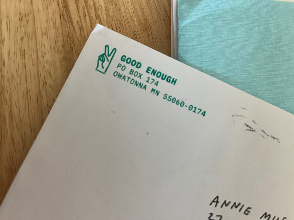 The corner of a white envelope with green peace fingers logo and address of Good Enough stamped in the return address and handwritten Annie visible on the address