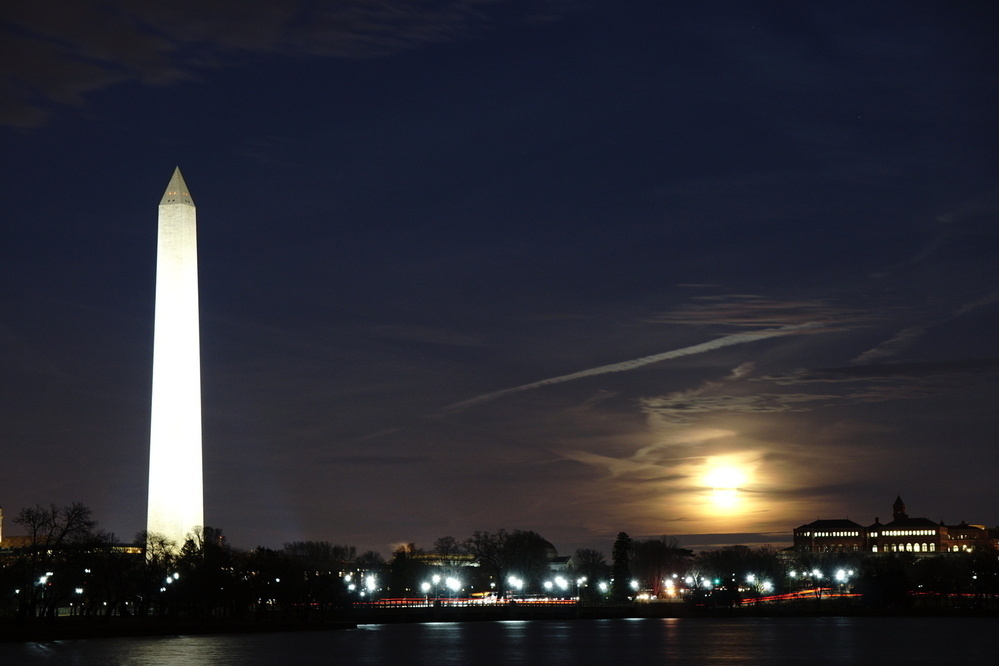 Night, long exposure photography of the Washington monument by the water on the left, and a full moon behind clouds. 