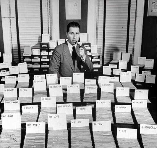 Mortimer J. Adler holding a pipe in his left hand and mouth posing in front of dozens of boxes of index cards with topic headwords including “law”, “love”, “life”, “sin”, “art”, “democracy”, “citizen”, “fate”, etc.