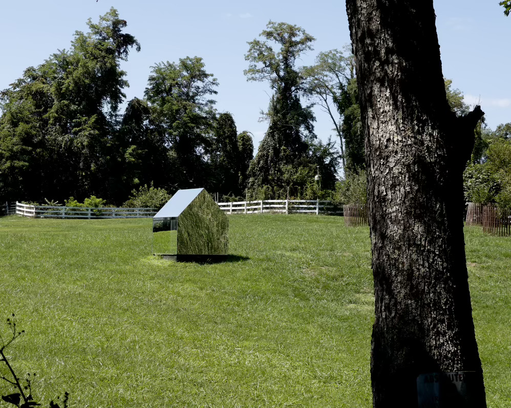 A small house shaped mirrored mausoleum sits in a small green grassy area in front of a white horse fence.