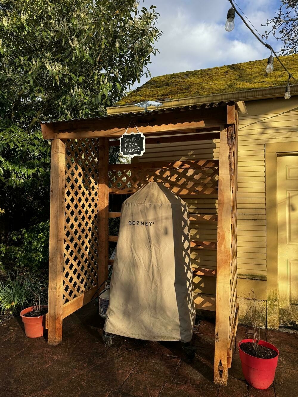 A wooden structure built to cover my outdoor pizza oven, with latticed sides and a corrugated tin roof with built-in ventilation. A small sign hangs from the top that says, in chalk, “David’s Pizza Palace”