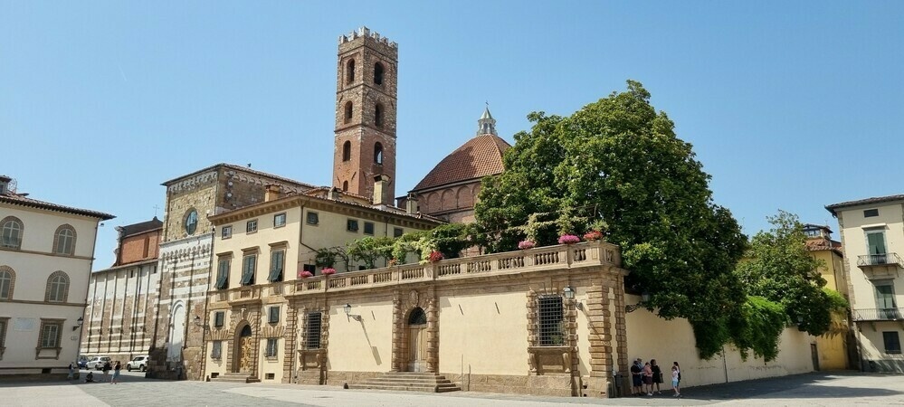 View from a piazza in Lucca, Italy. An old belltower in the background, a tree on a terraced building in front.