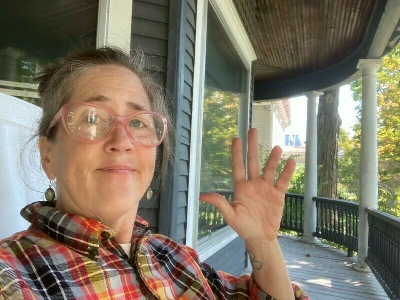 A 56 year old woman in an orange plaid shirt wearing pink glasses waves from her porch on a sunny day.