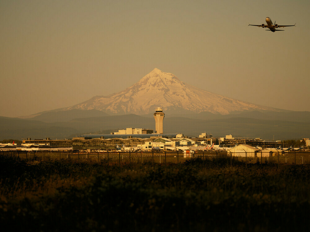 A photograph of Portland International Airport at golden hour. Mount Hood is centered in the background with the air traffic control tower. An Alaska Airlines plane is seen taking off from the runway headed in a westerly direction.