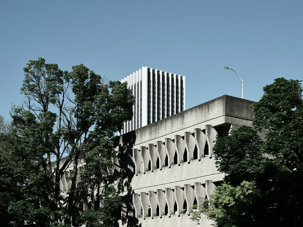 A parking garage in the brutalist architectural style with a skyscraper peeking from behind. A lone streetlamp post is on top of the parking garage.