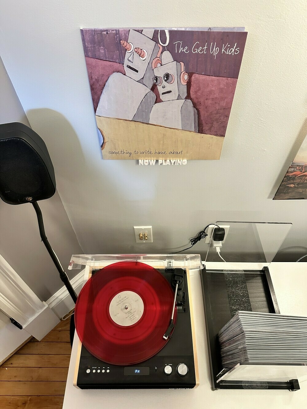 At the top of the photo - an album jacket perched on a “now playing” wall holder. At the bottom, a record player with the vinyl playing. To the left is a speaker and to the right are the tops of some stored comic books 