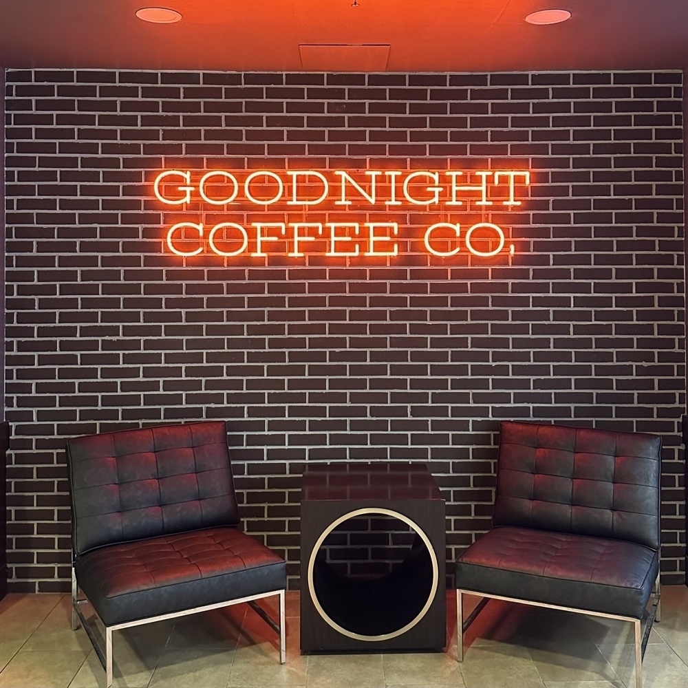 A brick wall with a neon sign that reads GOODNIGHT COFFEE CO is flanked by two black chairs with a small table in between.