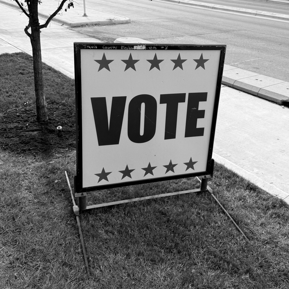 A sign with the word VOTE and stars around it is placed on a grassy area by the sidewalk.