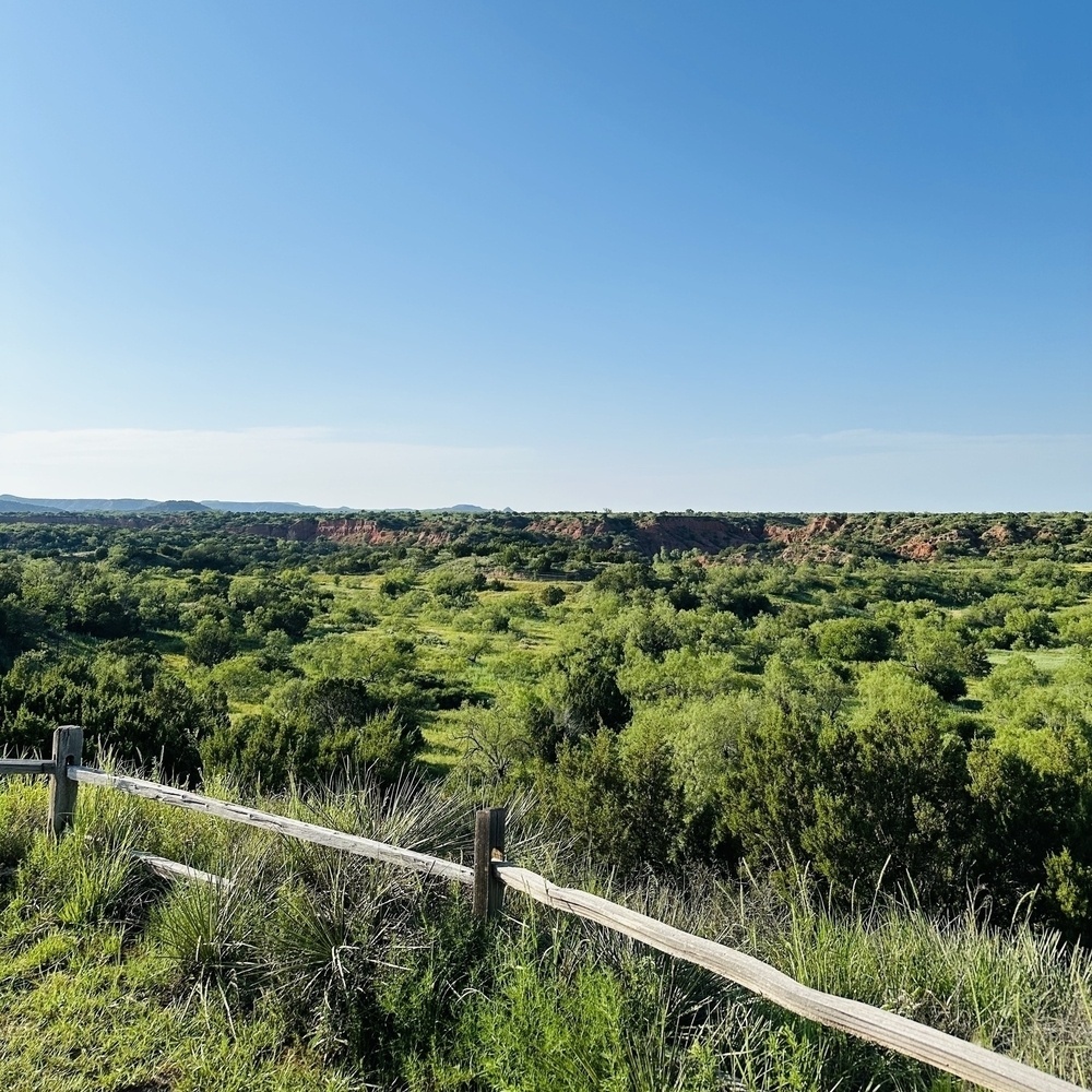 A scenic landscape features a wooden fence in the foreground overlooking a lush, green valley with distant red rock formations under a clear blue sky.