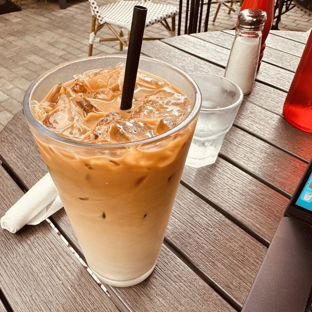 A glass of iced coffee with a straw sits on a wooden table alongside a glass of water, condiments, and a folded napkin in an outdoor setting.