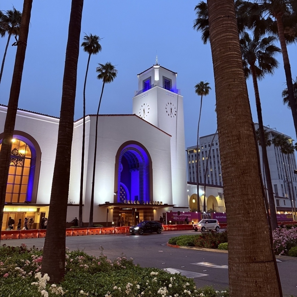 Union Station in Los Angeles is illuminated with purple and white lights, surrounded by tall palm trees.