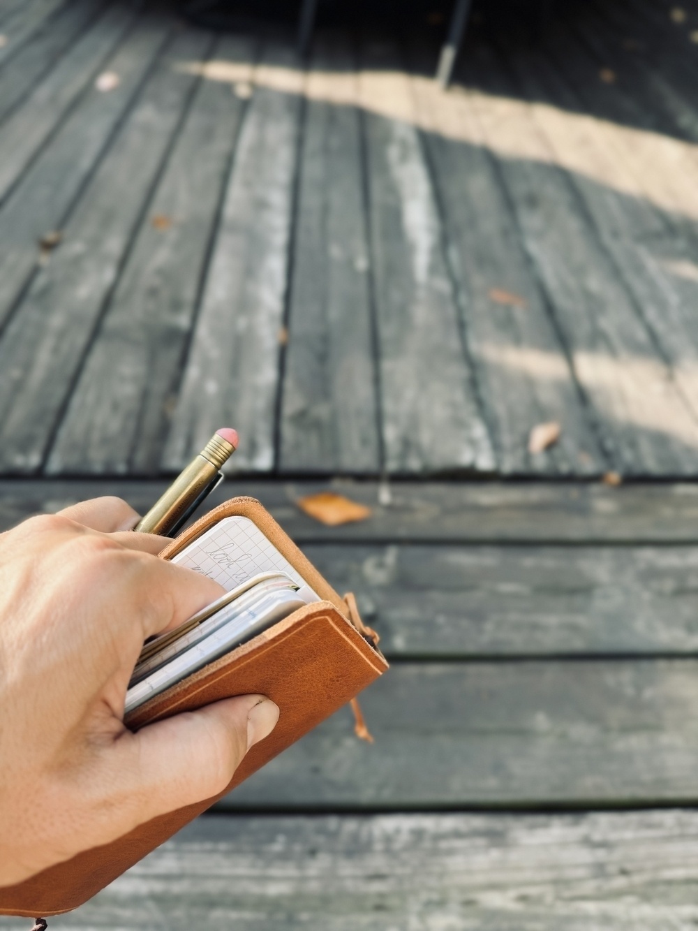 A hand holds a small leather notebook with a pencil tucked inside against a backdrop of wooden planks.