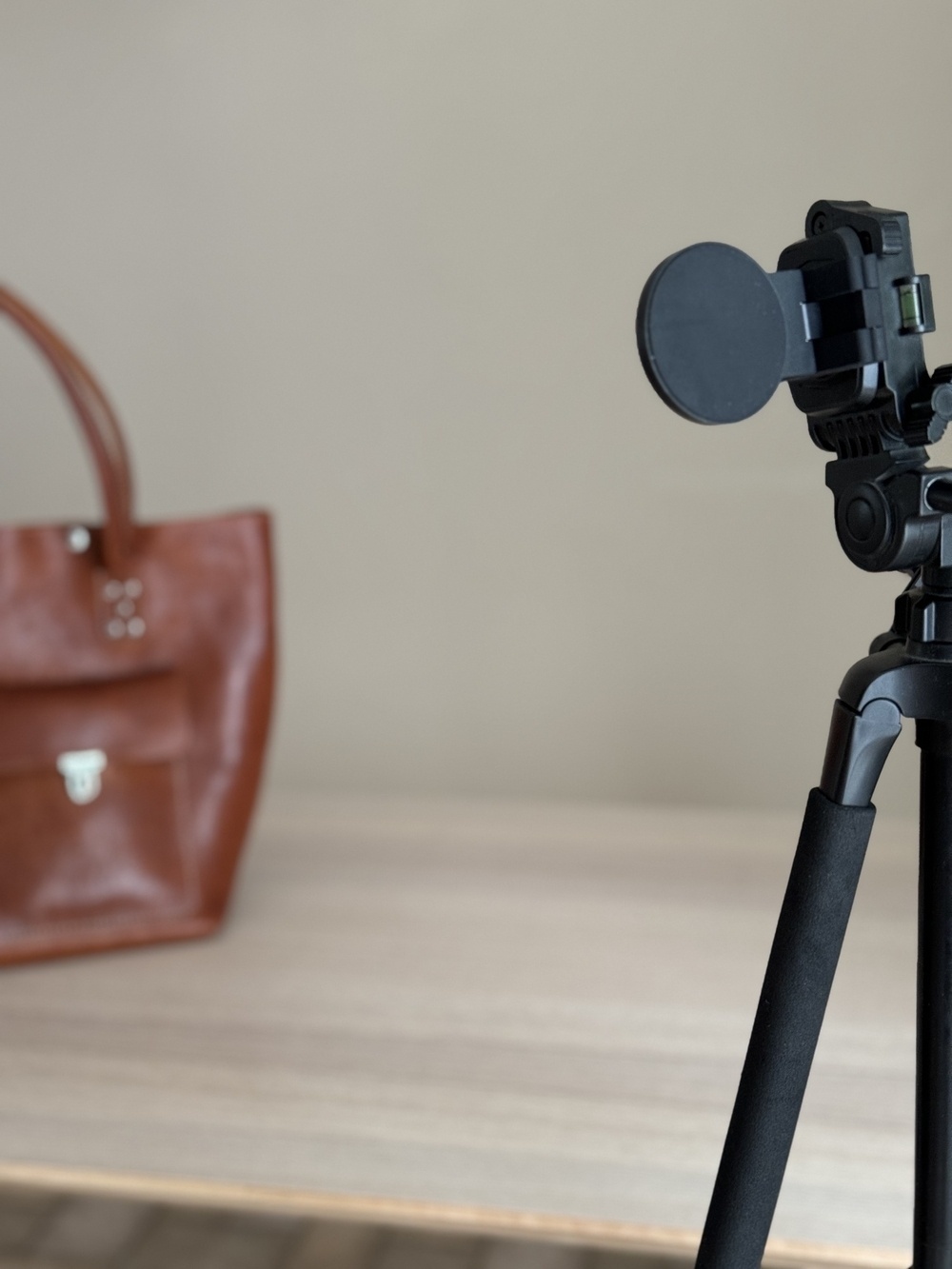 A camera tripod with a device mounted on it is set up indoors near a wooden table with a brown leather bag in the background.