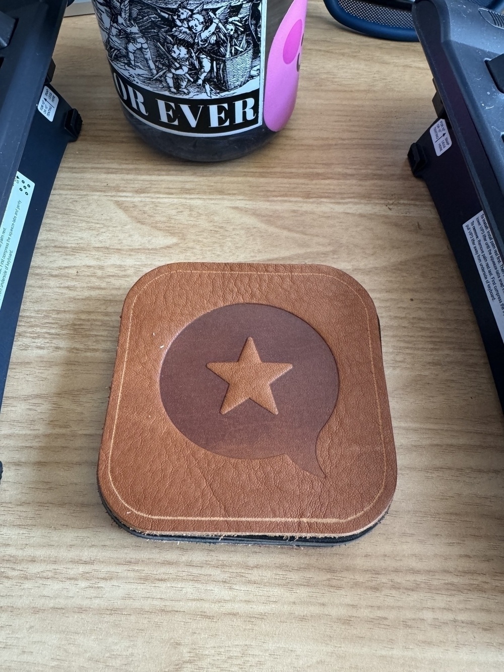 On a wooden surface, there's a leather coaster featuring a star within a speech bubble design (the micro.blog logo), flanked by two electronic devices, with a coffee mug in the background.