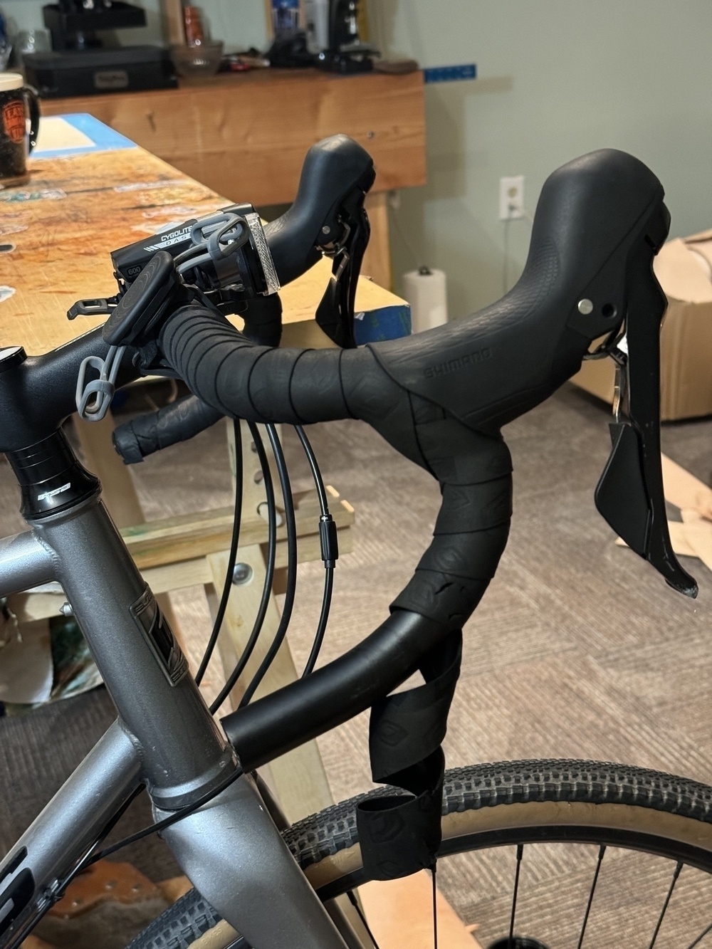 A close-up view of the handlebars and gear shifters of a road bicycle resting beside a wooden workbench.