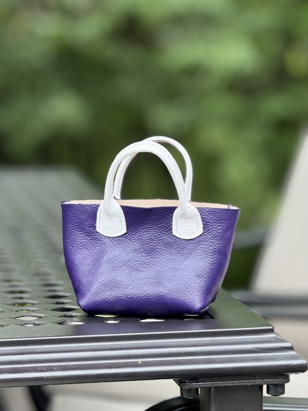 A small purple handbag with white handles is placed on a perforated metal table outdoors.