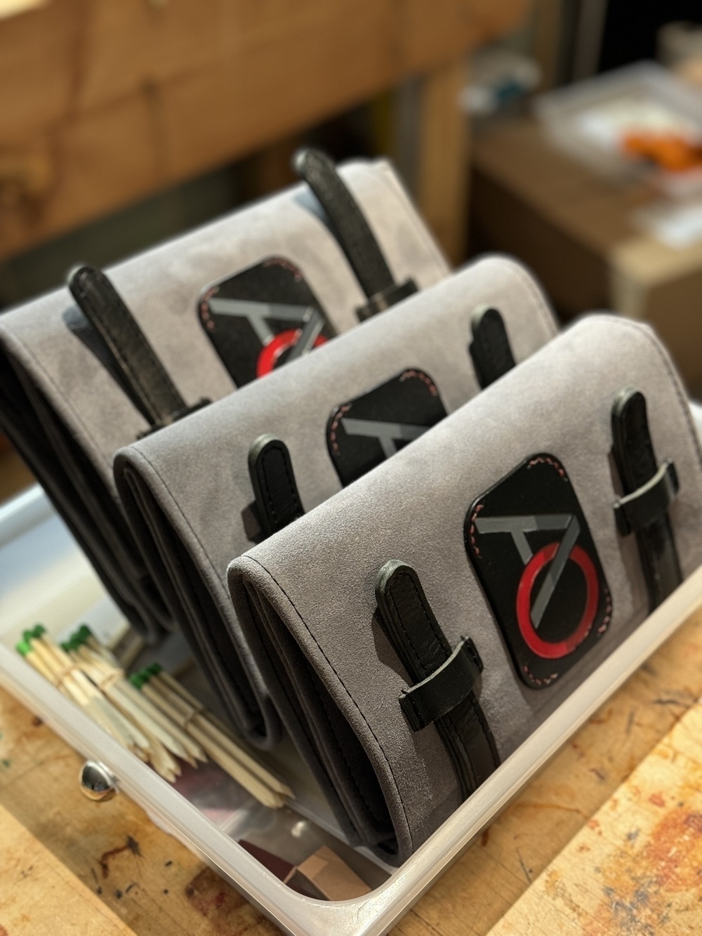 Three grey suede pouches with black straps and a logo featuring a red and black design are displayed in a container on a wooden surface.