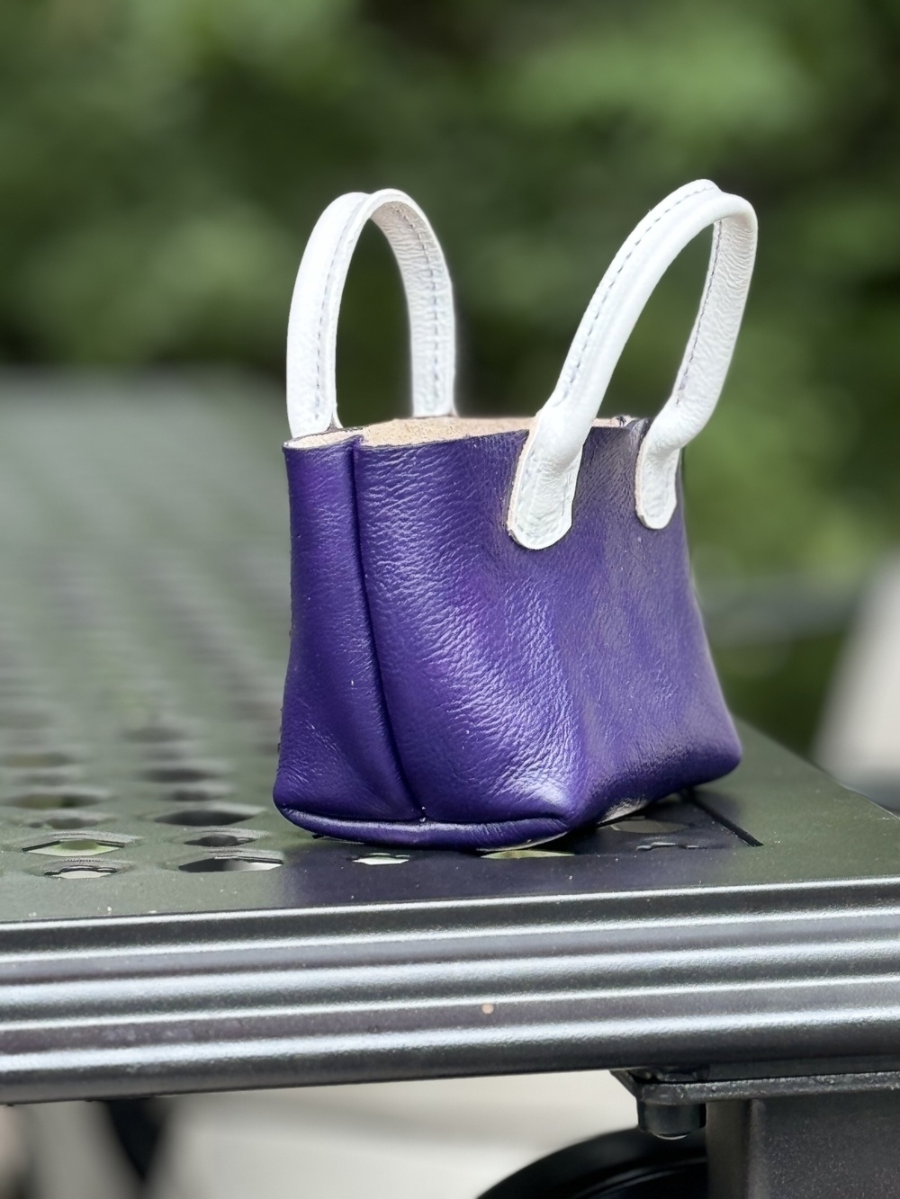 A small purple handbag with white handles is placed on a perforated metal surface outdoors.