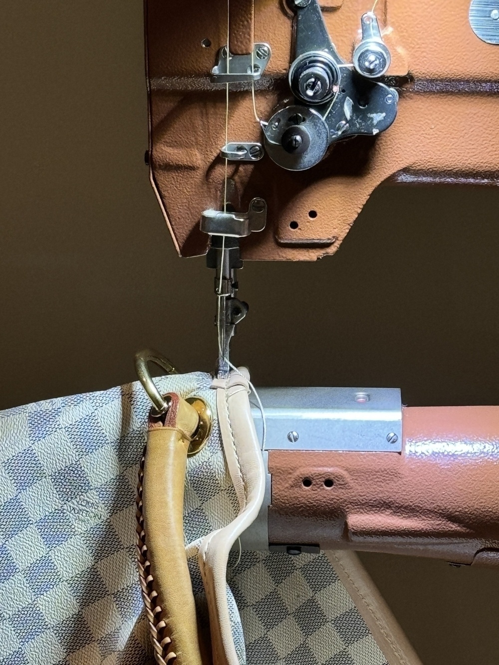 A sewing machine is stitching a beige leather handle onto a checkered fabric bag.