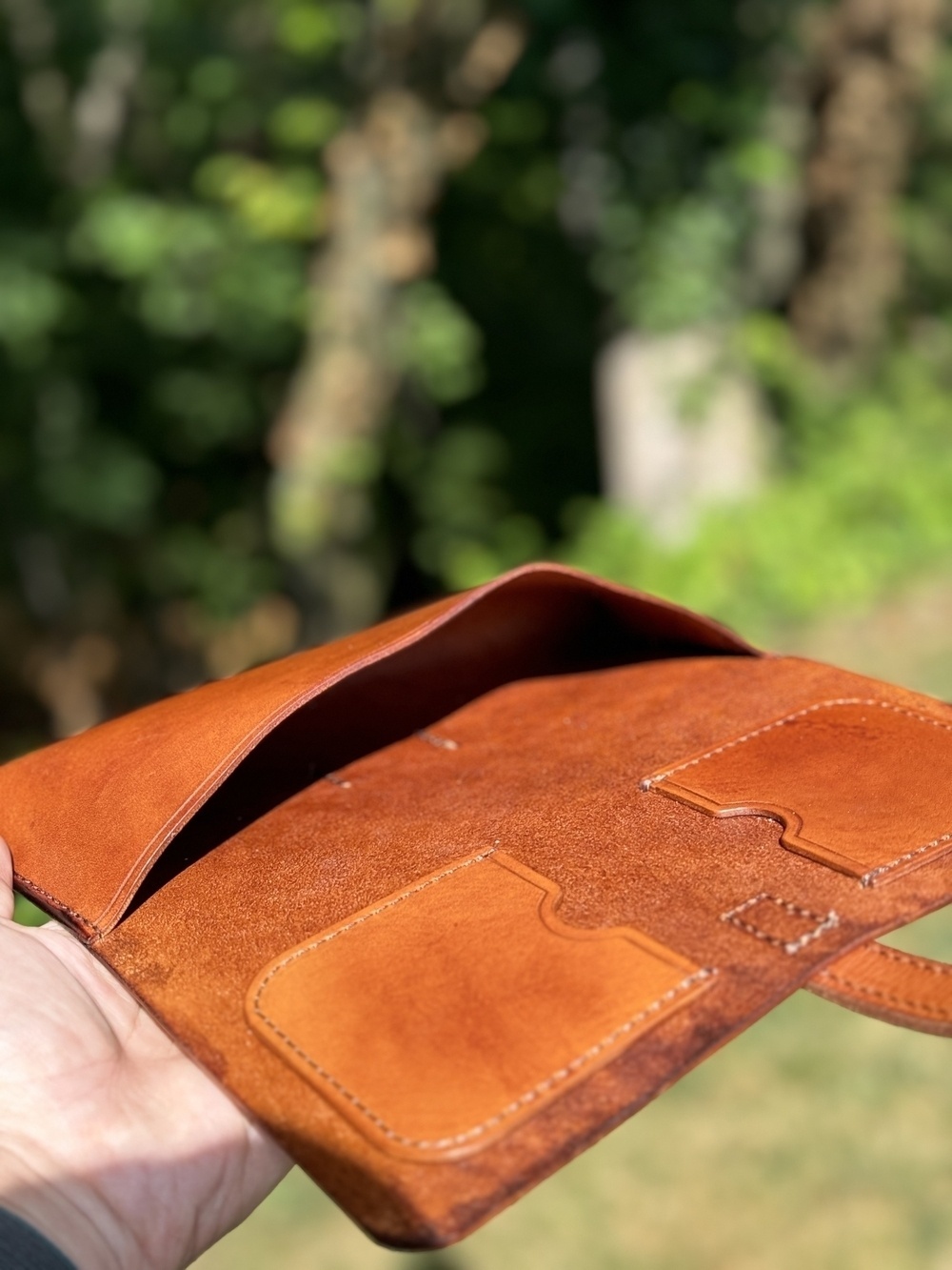 A hand holds an open, brown leather cigar case with cigar lighter and cigar cutter pockets and a large cigar pocket, against a blurred natural background.