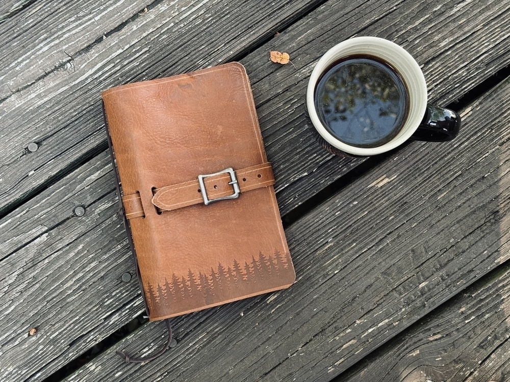 A leather-bound journal with a forest design and a buckle is placed next to a cup of coffee on a weathered wooden surface.