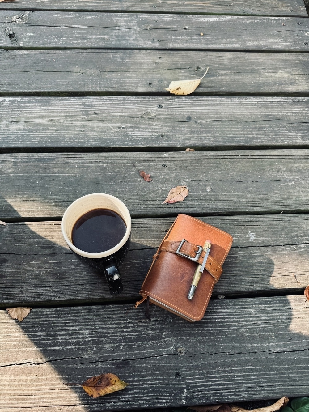 A cup of black coffee and a leather-bound notebook with a pen attached are placed on a wooden deck amidst scattered leaves.