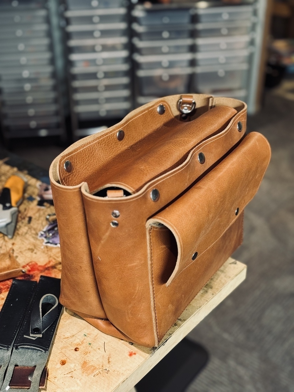 A tan leather bag in the process of being crafted is displayed on a workbench, with tools and materials around it and a background of organized storage drawers.