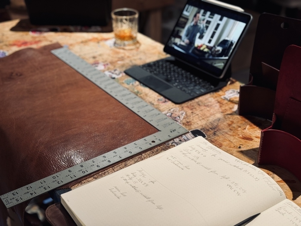 A workspace features a leather sheet being measured with a ruler, a notebook with handwritten notes, and a tablet playing a video (You’ve Got Mail).