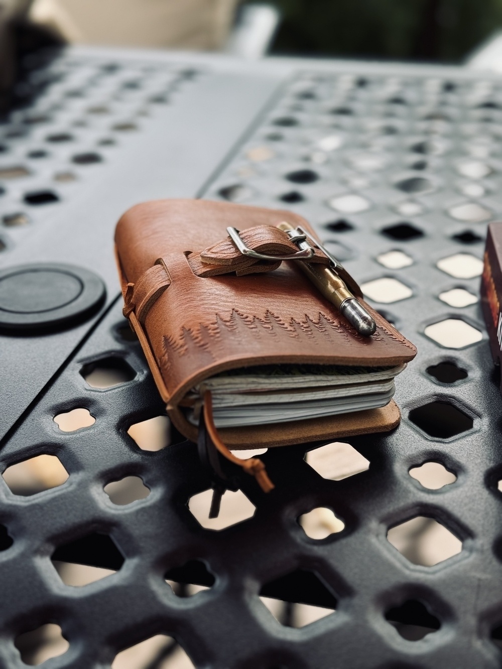 A leather-bound notebook with an embossed tree design and a pen attached is placed on a perforated metal table.