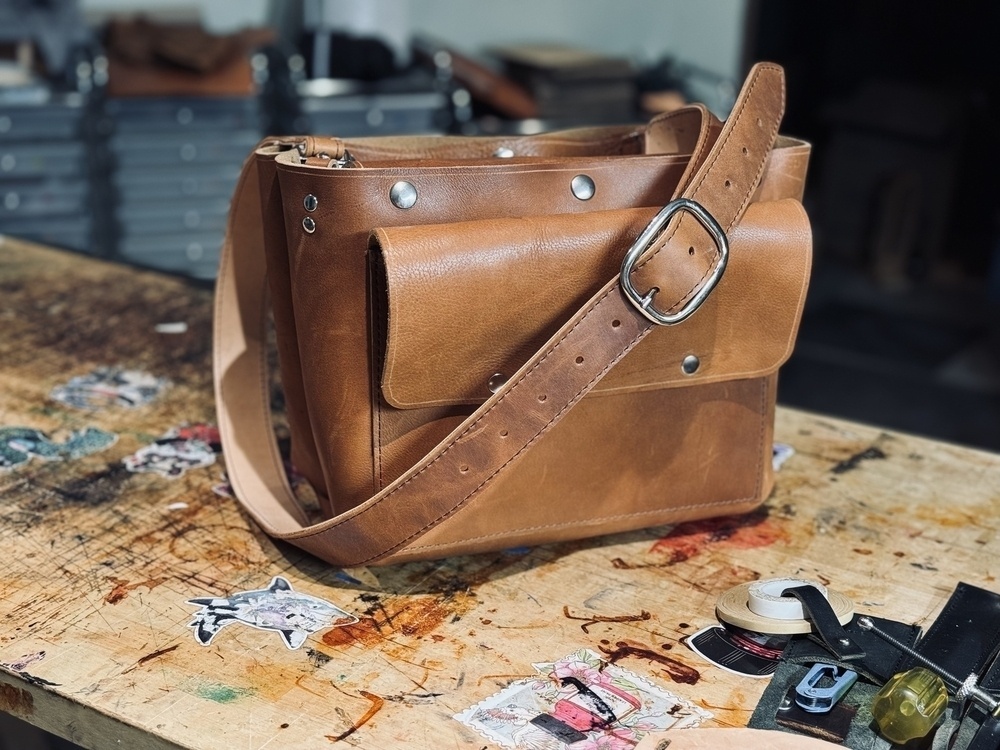 A leather shoulder bag with a large buckle is placed on a worn, cluttered workbench with various crafting supplies scattered around.