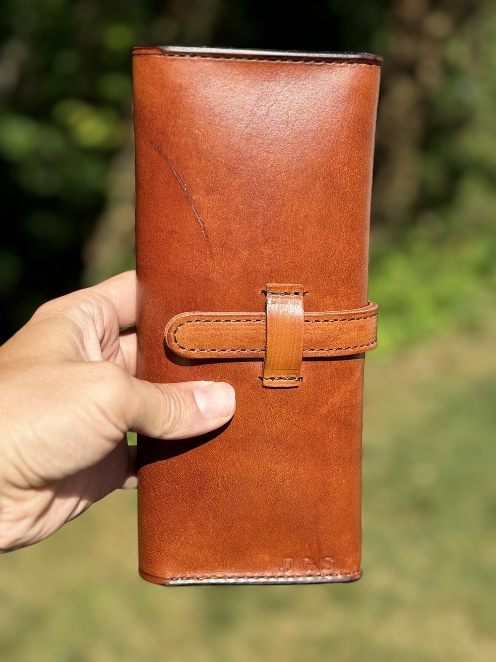 A hand is holding a brown leather cigar case outdoors, with a strap closure and green foliage in the background.