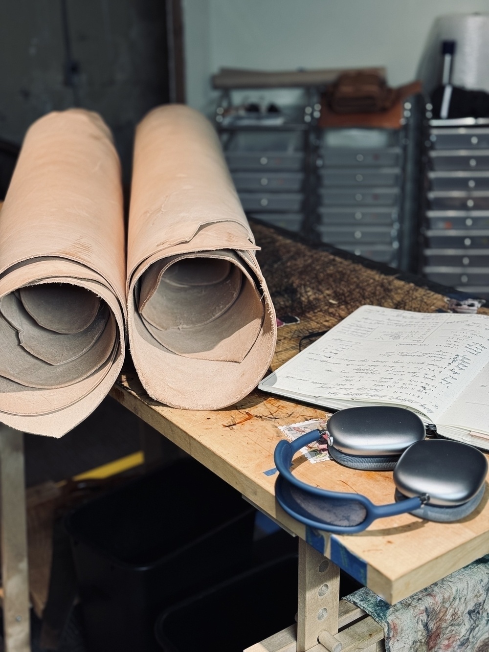 Two large rolls of leather are on a workbench beside a notebook and headphones, with storage drawers in the background.