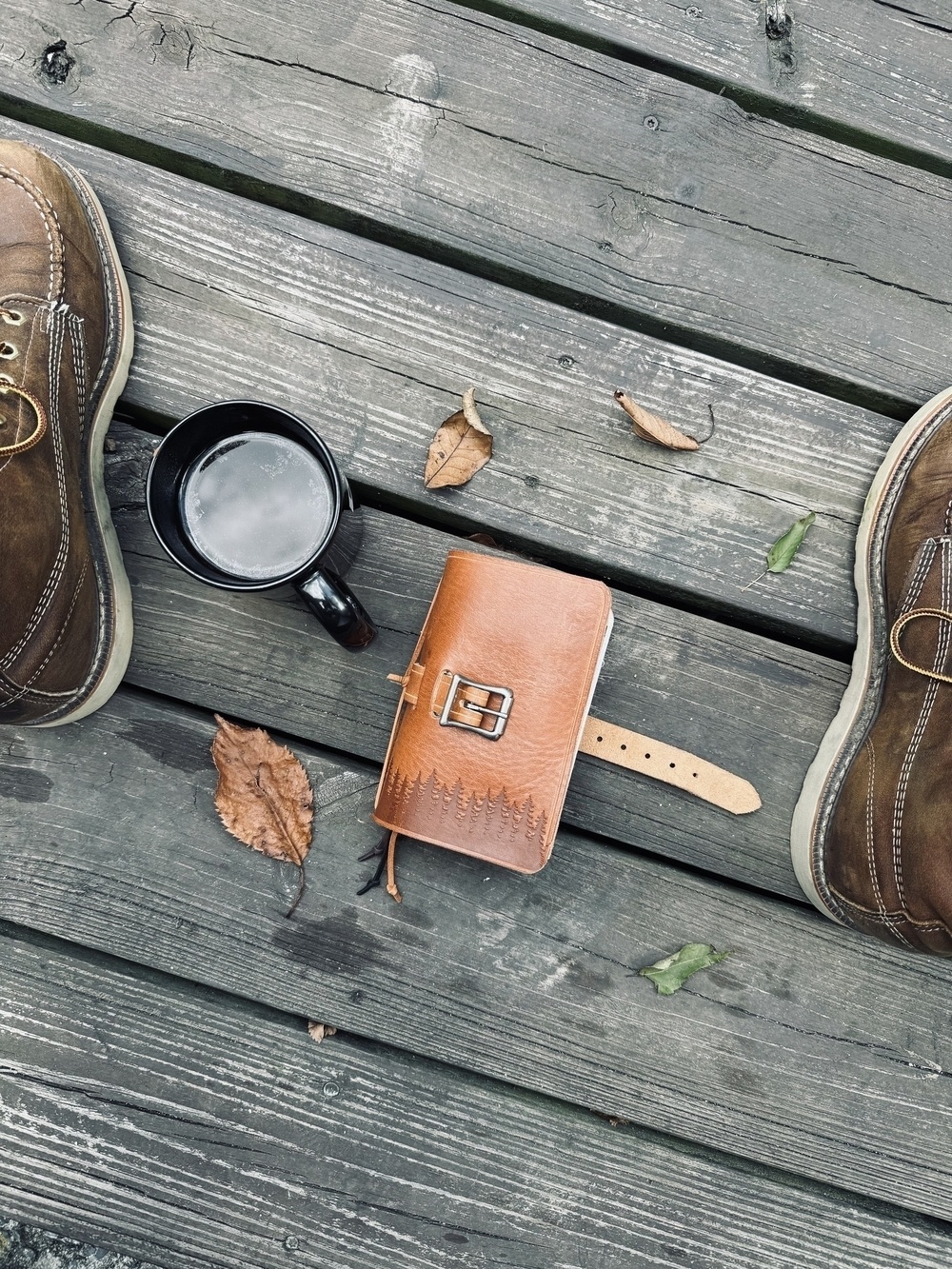 A pair of brown boots, a black mug, fallen leaves, and a small leather-bound journal with a buckle lie on a wooden surface.