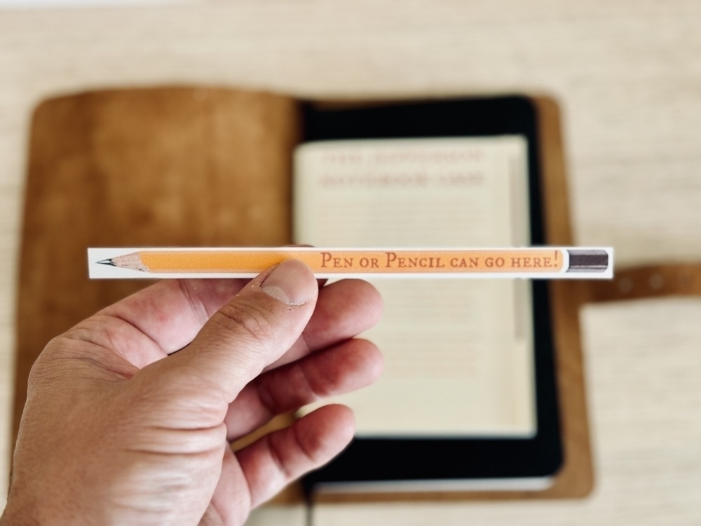 A hand is holding up a pencil with the words "Pen or Pencil can go here!" in front of an open notebook with a leather cover.