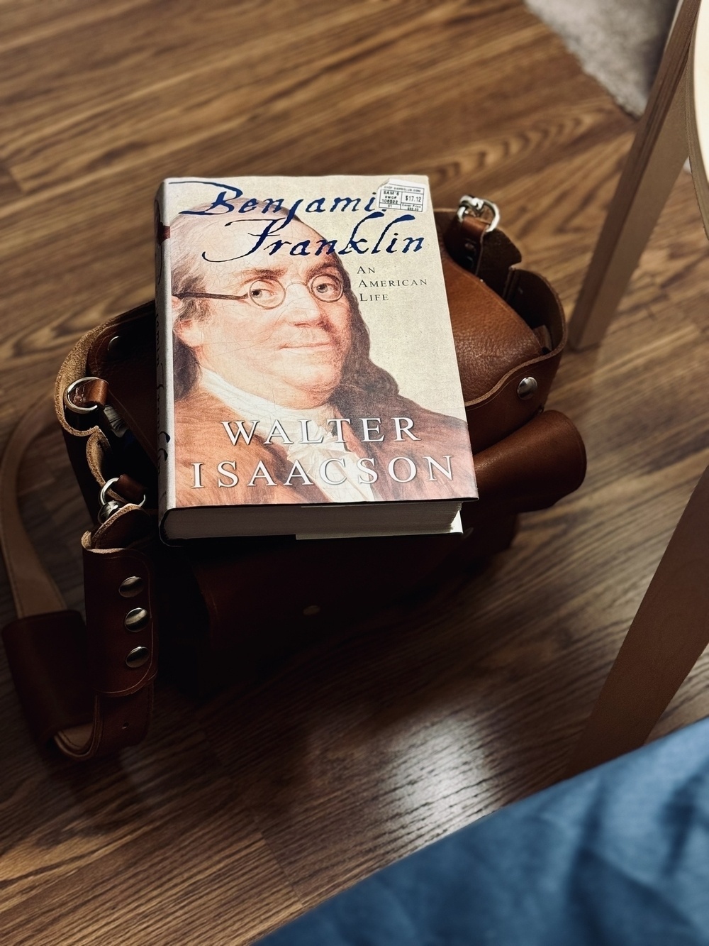 A book titled "Benjamin Franklin: An American Life" by Walter Isaacson is placed on top of a brown leather bag on a wooden floor.