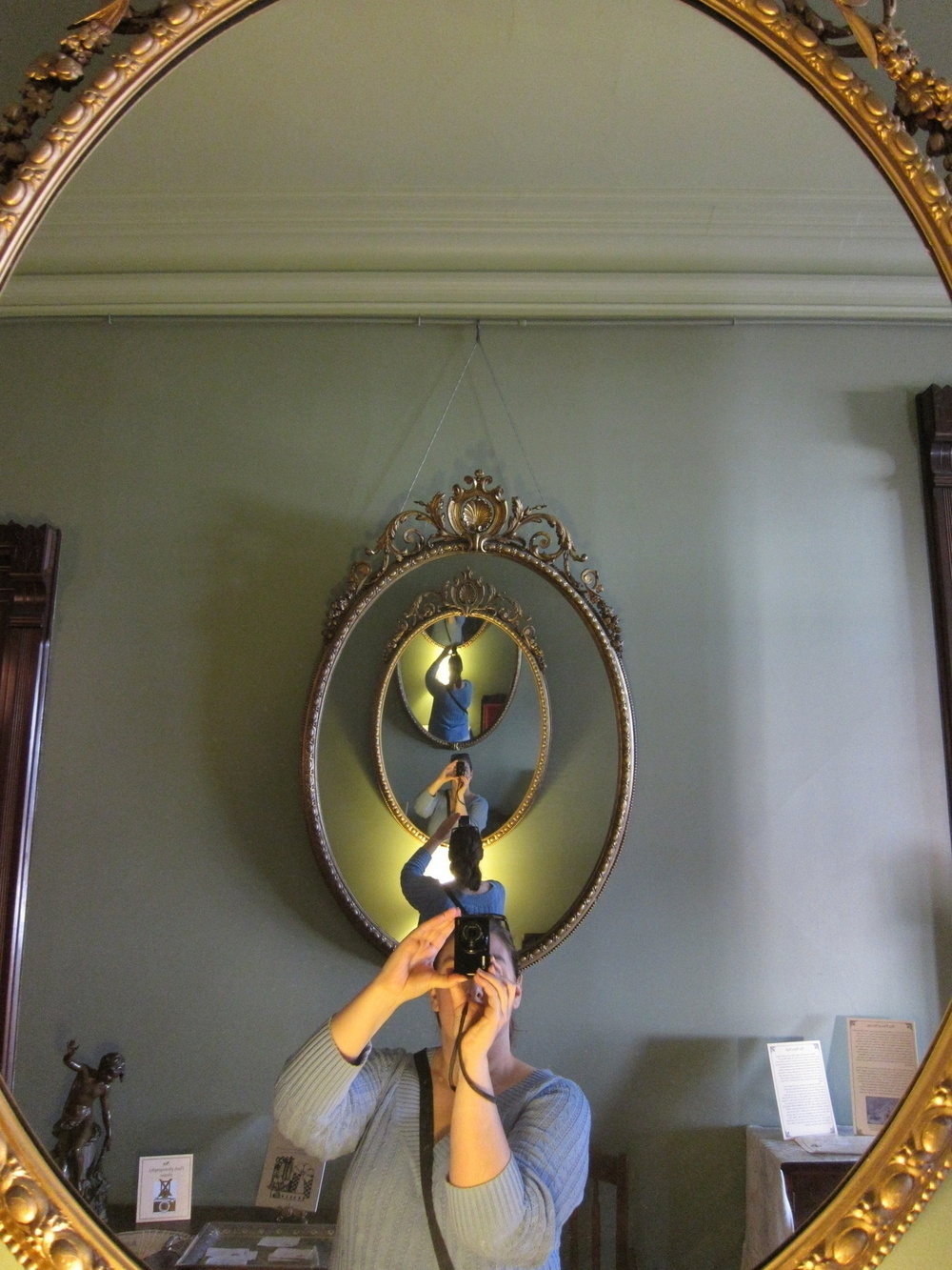 Opposing mirrors on walls create a repeating front-to-back self portrait of the photographer: a woman in a blue sweater with long hair pulled up in the back holding a small black camera in front of her face.