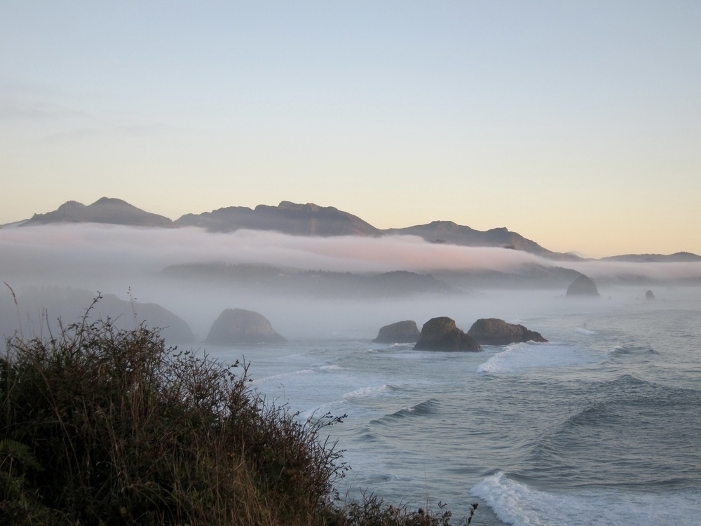 Standing on a coastal headland looking toward Cannon Beach near sunset, a pink fog flows from the valleys and along the beach and coastline. The whole scene has a pink and dreamy quality.