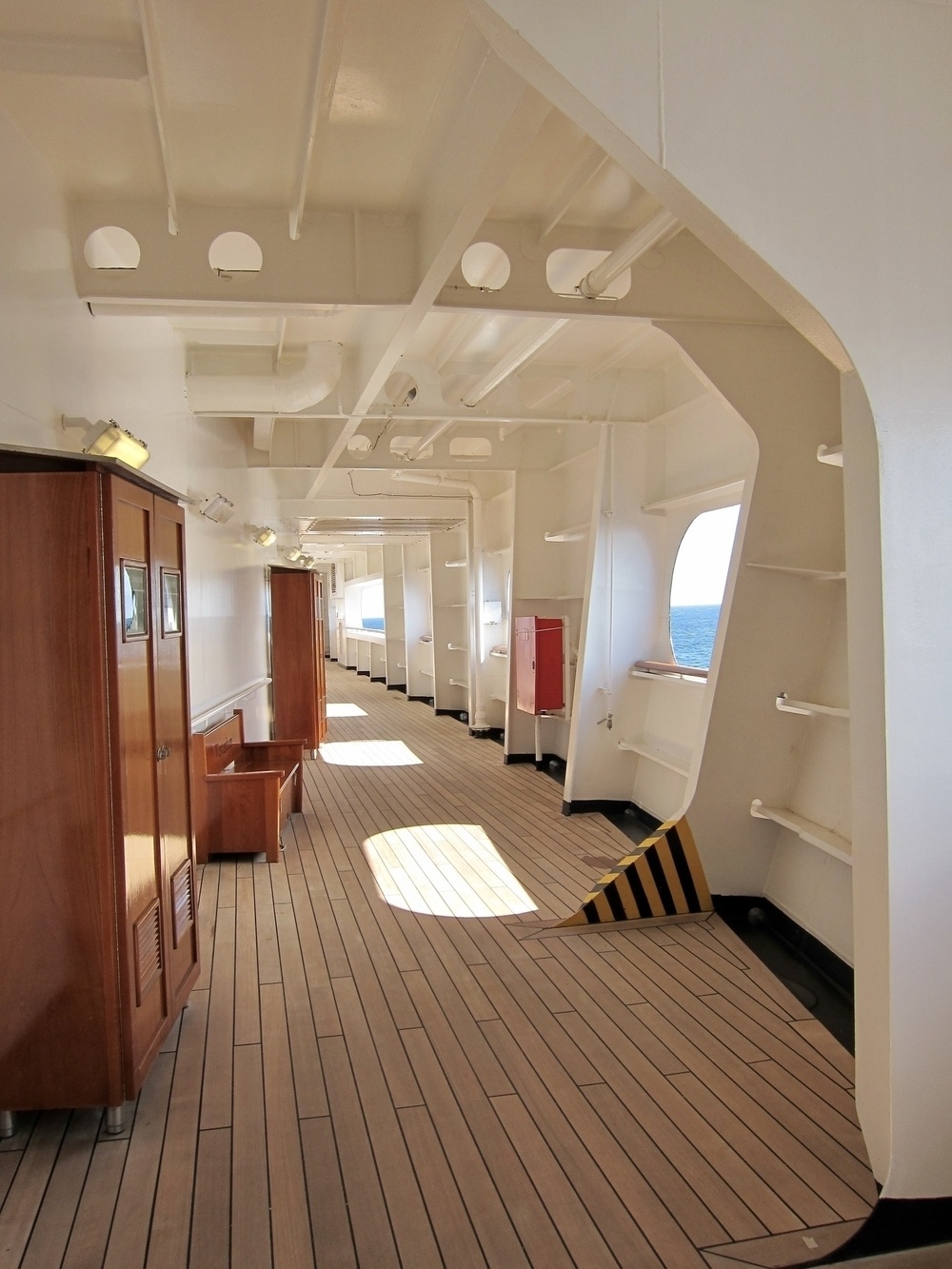 On the covered deck of a cruise ship, white painted metal arched supports and openings in the side of the ship reveal sun shining onto the wood deck creating large sun spots, along with punches of color from a warning strip with yellow and black, the warm wood of two cabinets and a bench on the inner wall, and a red metal box attached to the outer wall.