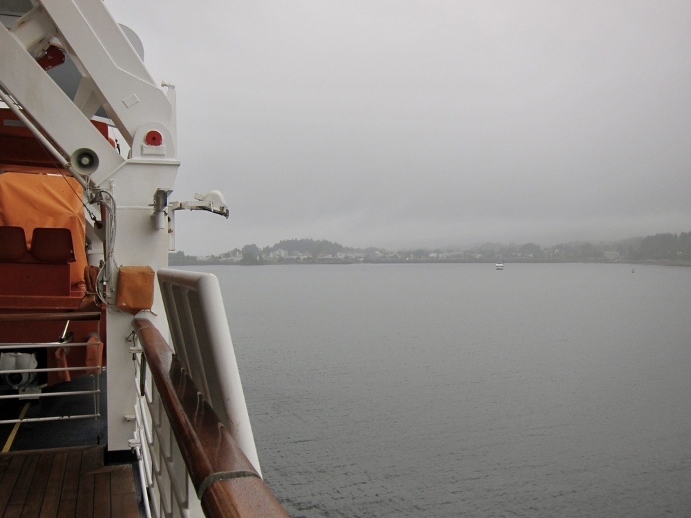 Looking out over water from the deck of a ship, part of a railing and other ship infrastructure visible on the far left of the picture, the view over the water shrouded by mist and fog with land and a single small boat in the distance.