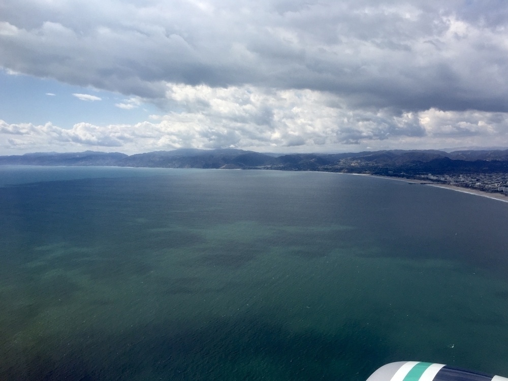 A glimpse of an airplane engine in the lower right gives way to the blue and green of water off the coast of Los Angeles from the air, with the coastline to the north arching in the distance.