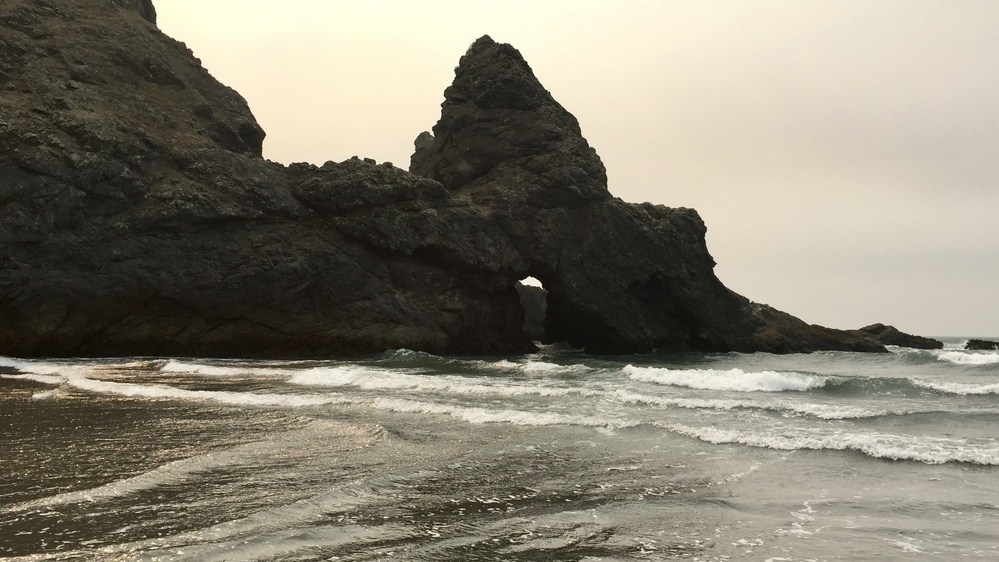 Under a slightly broody overcast sky, waves lap the shoreline and large craggy basalt seastacks rise with a very large pointed arch directly in front.