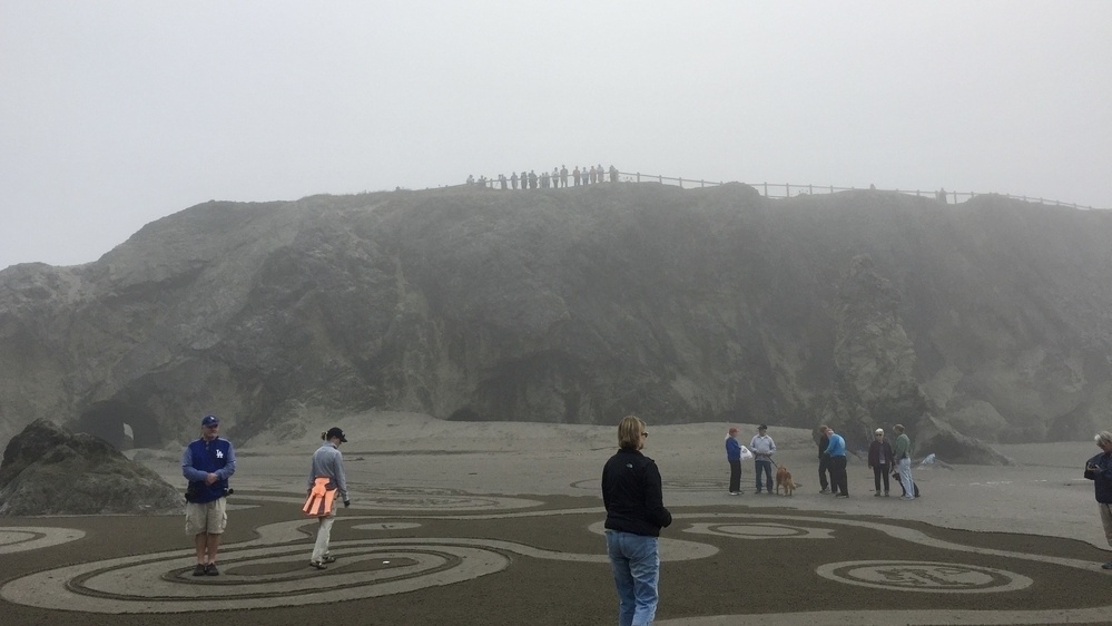 Standing on a beach looking toward a large rock outcropping: people mill about on a sand labyrinth in the foreground, and on top of the outcropping stand a line of people watching, appearing as silhouettes in the fog.