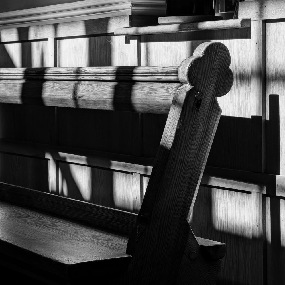 Sunlight through a window throws shadows over a wooden bench, in black and white