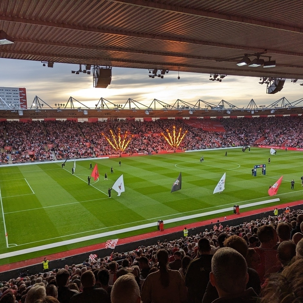 View across the pitch of St Mary’s Stadium. Fan shaped jets of flame light up in front of the stands on the far side of the pitch.