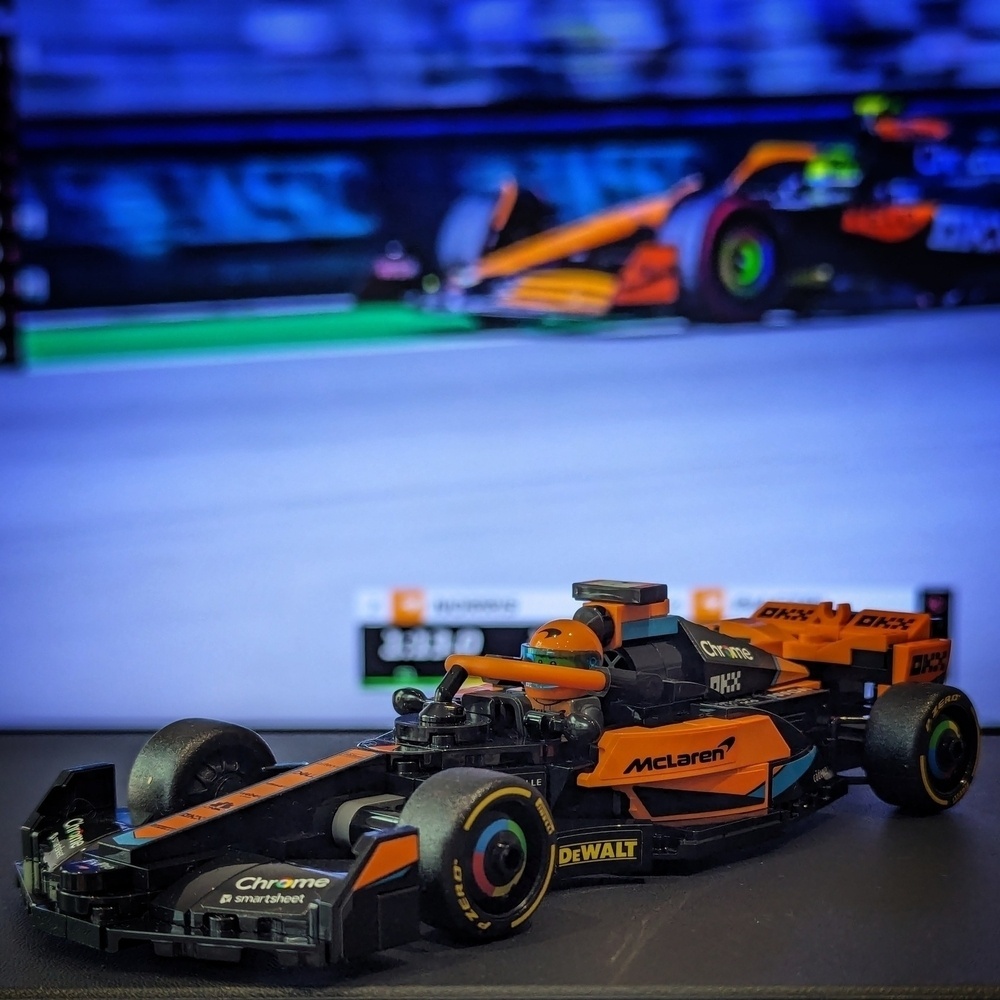 A Lego Speed Champions Formula 1 papaya McLaren sits on a black surface in front of qualifying from Imola being shown on a soft-focus TV screen behind.