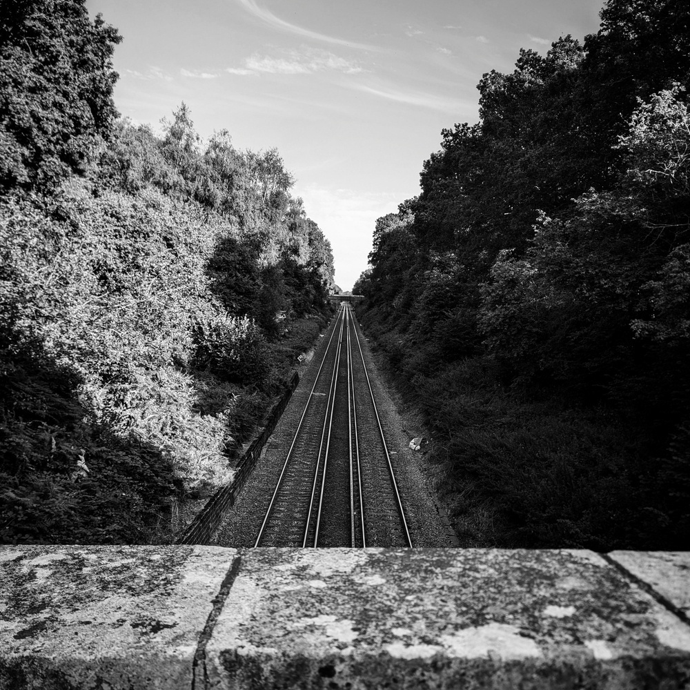 Black and white image of straight train tracks receding into the centre of the frame. The tracks are flanked by banks of foliage and larges stone bricks of a bridge wall are partially visible in the foreground.