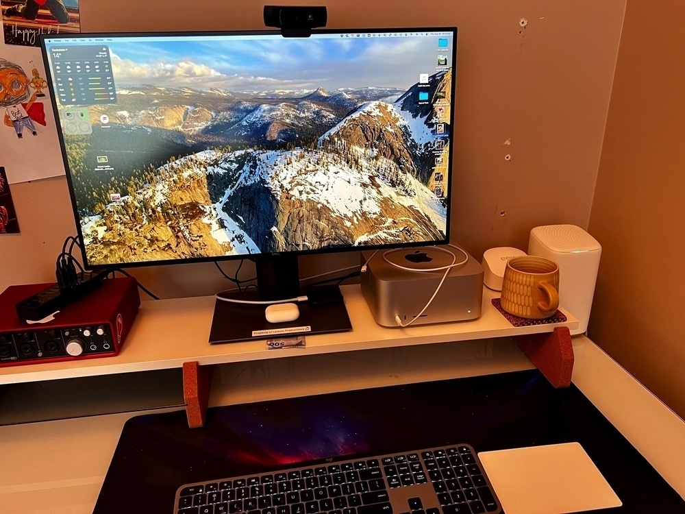 Photo of a computer desk with a keyboard, Magic Trackpad on it, with a desk riser shelf holding a monitor, Mac Studio, coffee mug, and focusrite 4 track recording preamp.